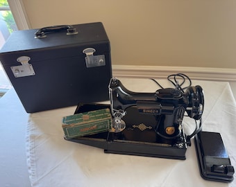 May include: A black Singer sewing machine with a matching black carrying case, a black foot pedal, and a small green rectangular box labeled Singer. The machine features gold-tone decorative accents on the base and a power cord attached.