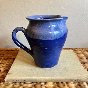 May include: A ceramic pitcher featuring a two-tone blue glaze with a lighter, speckled blue upper section and a solid, deep blue lower section. The pitcher has a curved handle on the left side and a wide, rounded spout at the top rim.