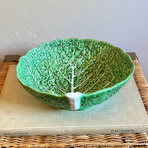 May include: A green ceramic bowl shaped like a cabbage leaf with a textured surface and a white stem detail at the base. The bowl features a scalloped rim and a glossy finish, mimicking the appearance of a fresh head of cabbage in a round form.