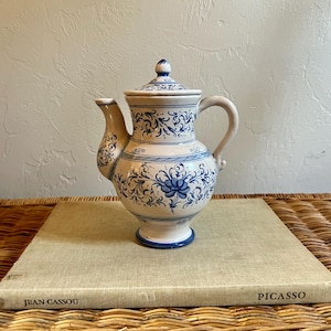 May include: A ceramic teapot with a lid, featuring a cream-colored base decorated with intricate blue floral and scroll patterns. The teapot has a curved handle, a spout, and a pedestal base, all accented with blue trim and painted details.