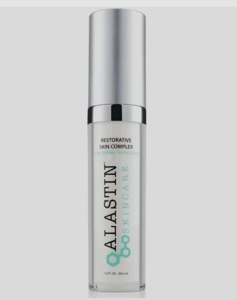 Op de afbeelding: Een flesje huidverzorgingsproduct met een zilveren dop en een transparante body. Het etiket leest "ALASTIN Skincare" met de woorden "Restorative Skin Complex" en "1.0 fl oz. 29,6 ml". De achtergrond is lichtgrijs.