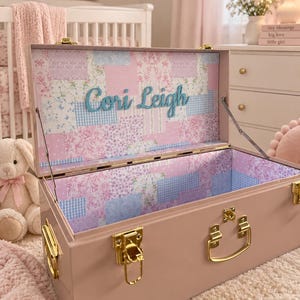 May include: A dusty pink metal storage trunk with gold-toned latches and handles. The interior lid and walls are lined with a patchwork pattern of floral and gingham prints in shades of pink, blue, and white, featuring the name Cori Leigh.