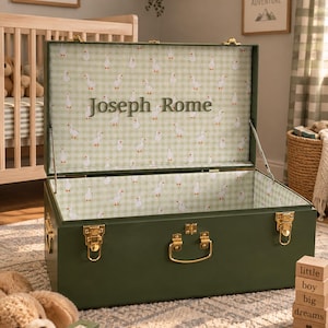 May include: A rectangular olive green storage trunk with gold-toned metal latches and a handle. The interior is lined with a light green and white gingham fabric featuring a repeating goose pattern and the personalized text Joseph Rome.