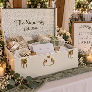 May include: A white rectangular storage chest with gold latches and handle, featuring the text The Sinacoris Est. 2026 inside the lid. The chest is filled with wrapped gifts and envelopes, accompanied by a sign reading GIFTS and CARDS.