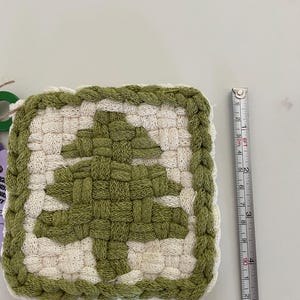 May include: A square woven potholder featuring a green tree design on a cream background with a braided green border. A measuring tape placed alongside shows the item measures approximately 4.5 inches in height and width.