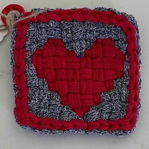 May include: A square woven coaster featuring a red heart design centered against a background of speckled black and white yarn. The border is constructed from thick red yarn, and a small red ring is attached to the corner with a piece of twine.