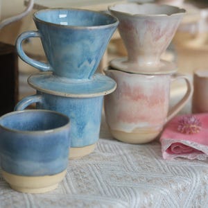 May include: A collection of ceramic coffee drippers and mugs featuring a two-tone design with a smooth, unglazed beige base and a glossy, reactive glaze in shades of sky blue or soft pink. The pieces have rounded edges and sturdy handles.