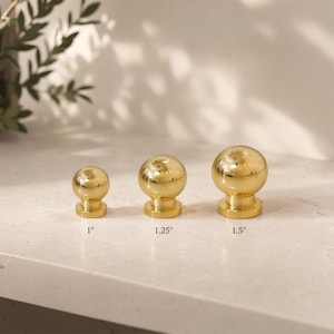 May include: Three polished gold-tone spherical cabinet knobs arranged in a row. The knobs are labeled with their respective diameters of 1 inch, 1.25 inches, and 1.5 inches, showing a smooth, reflective metallic finish and a circular base.