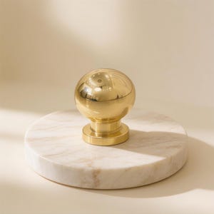 May include: A polished gold-toned spherical cabinet knob with a smooth, reflective surface and a cylindrical base, mounted on a circular white marble disc featuring subtle grey veining throughout its flat, polished surface.