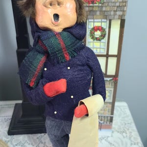 May include: A figurine of a person singing, wearing a red knit cap, a dark blue sweater with small white dots, a green and red plaid scarf, and red mittens. The figure holds a long, blank, light-colored paper scroll and wears dark gray trousers.