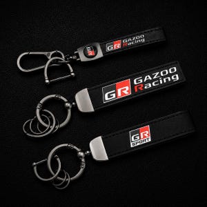 Puede incluir: Tres llaveros negros con herrajes plateados. Cada llavero presenta una correa negra con el logotipo "GR" en rojo y blanco, y las palabras "GAZOO Racing" o "SPORT". Los llaveros están sobre una superficie negra.