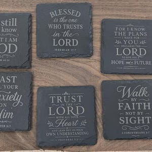 May include: A set of nine square black slate coasters with rough, natural edges. Each coaster features engraved white text with various religious verses and inspirational quotes, including phrases like Peace I leave with you and Trust in the Lord.