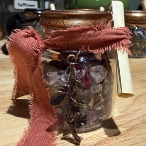 May include: A clear glass jar with a metallic bronze lid, tied with a frayed terracotta-coloured ribbon. Inside are clear and purple crystal chips and dried botanicals. A bronze-toned metal charm shaped like a branch with leaves hangs from the neck.