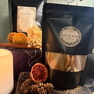 May include: Two black pouches labelled Into The Woods Simmer Pot containing dried orange slices, spices, and botanicals. The front pouch features a circular label with text, a clear window showing contents, dried orange slices, and small pinecones.
