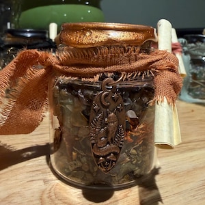 May include: A clear glass jar filled with small dark stones and dried botanical elements. The lid is sealed with metallic bronze wax. A frayed orange fabric ribbon is tied around the neck, holding a bronze-toned oval charm featuring a fox.