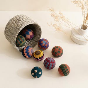May include: A gray woven basket tipped over, spilling out several colorful, patterned crochet balls. The balls feature various designs in shades of blue, orange, green, and purple. A white vase with dried flowers is in the background.