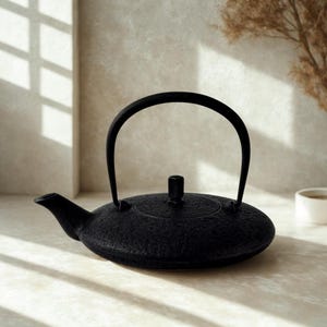 May include: A matte black cast iron teapot with a low, wide, circular body and a textured surface. It features a curved spout, a tall arched handle, and a small, cylindrical lid knob centred on the flat top of the vessel.