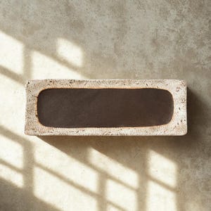 Puede incluir: Una bandeja rectangular con una base interior lisa de color marrón oscuro y un borde ancho, texturizado y de color beige claro. El borde exterior presenta una superficie rugosa y porosa con variaciones naturales de color.