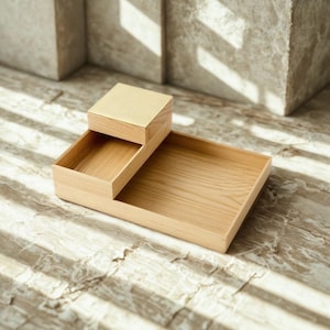 Op de afbeelding: Een bureau-organizer van lichtgekleurd hout met een grote rechthoekige basisplaat en een kleiner, verhoogd vierkant vak aan één zijde. De natuurlijke houtnerf is zichtbaar over de gehele structuur, die strakke en rechte randen heeft.
