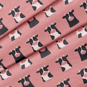May include: A fabric featuring a repeating pattern of stylized cow heads on a dusty rose background. Each cow head is rendered in black and white with a pink snout and small horns, arranged in a consistent grid-like layout across the textile surface.