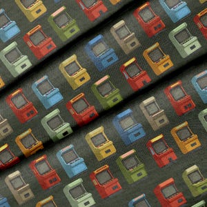 May include: A dark gray fabric featuring a repeating pattern of miniature arcade cabinets in various colors including red, blue, yellow, green, and beige. Each cabinet has a screen, control panel, and joystick, arranged in a diagonal grid layout.