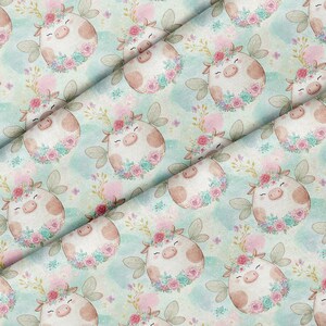 May include: A fabric pattern featuring repeating illustrations of round, light brown pigs with closed eyes and smiling expressions. Each pig is adorned with a floral crown of pink and teal roses and green leaves against a soft, light blue background.