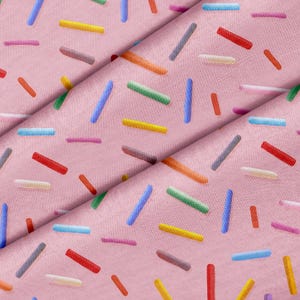 Könnte beinhalten: Ein hellrosa Stoff mit einem sich wiederholenden Muster aus kleinen, zylindrischen Streuseln in verschiedenen Farben wie Blau, Gelb, Grün, Rot, Orange und Weiß, die verstreut und in unterschiedlichen Richtungen auf der Textiloberfläche angeordnet sind.