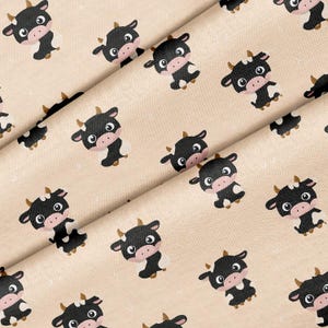 May include: A fabric featuring a repeating pattern of cartoon cows on a light beige background. Each cow has a black body, a pink snout, and small brown horns. The cows are arranged in a diagonal orientation across the textile surface.