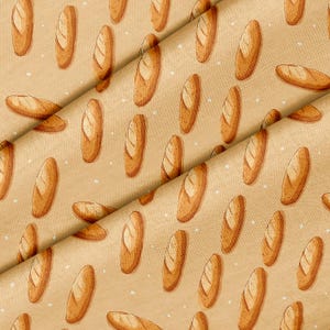 May include: A tan fabric featuring a repeating pattern of golden-brown baguette illustrations. The bread loaves are oriented in various directions across the surface, interspersed with small, scattered white dots that resemble flour particles.