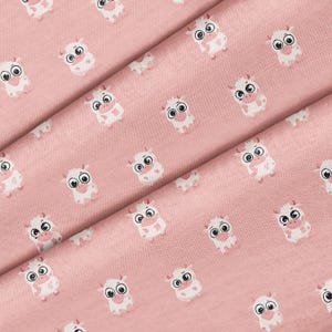 May include: A light pink fabric featuring a repeating pattern of small, white cartoon cows with large, expressive eyes and pink snouts. The textile has a visible woven texture and the cow illustrations are evenly spaced across the entire surface.