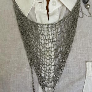 May include: A silver-toned multi-strand chain necklace featuring a wide, triangular mesh design that drapes over the chest. The necklace includes several dangling silver chains and dark grey spherical beads at the bottom and side accents.