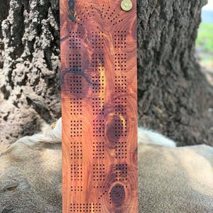 May include: A rectangular wooden cribbage board with numerous drilled holes for peg placement. The board is made of reddish-brown wood with a natural grain pattern and a decorative brass accent. The board is designed for the classic card game.