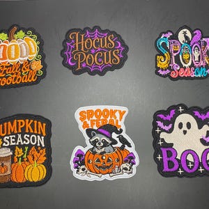 May include: Six embroidered patches featuring Halloween and autumn themes. Designs include a football pumpkin, Hocus Pocus text with spiderwebs, Spooky Season text, a coffee cup with pumpkins, a raccoon in a pumpkin, and a ghost with bats.