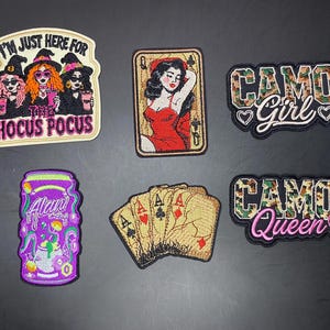 May include: Six embroidered patches featuring various designs: three witches with the text I'M JUST HERE FOR THE HOCUS POCUS, a Queen of Spades playing card, a CAMO GIRL patch, a purple Alani energy drink can, four playing cards, and a CAMO QUEEN patch.