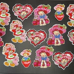 May include: A collection of embroidered patches featuring the character Strawberry Shortcake in various poses, including sitting on a red suitcase, standing with a pink cat, and framed within red heart shapes, all with pink, red, and green colors.