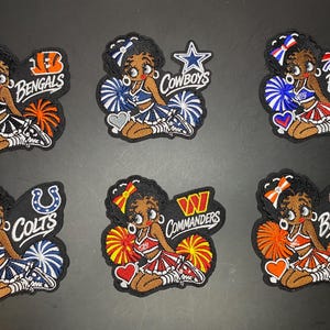 May include: Six embroidered patches featuring the cartoon character Betty Boop dressed as a cheerleader. Each patch displays a different NFL team name and logo, including the Bengals, Cowboys, Bills, Colts, Commanders, and Browns, with colorful pom-poms.