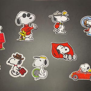 May include: A collection of nine embroidered patches featuring the character Snoopy in various outfits and activities, including wearing sunglasses, a red sweater with the text Joe Cool, a hat, a space helmet, and driving a red convertible car.