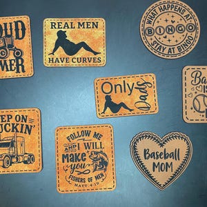 May include: A collection of seven tan faux leather patches with black laser-engraved designs. The patches feature various shapes including squares, rectangles, a circle, and a heart, displaying text and graphics related to farming, baseball, and fishing.