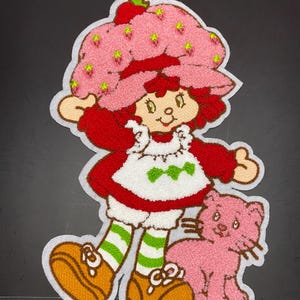 Peut inclure: Un écusson en tissu représentant un personnage de dessin animé aux cheveux roux portant un chapeau rose à motifs de fraises, une veste rouge, un tablier blanc avec des feuilles vertes, des chaussettes rayées vert et blanc et des chaussures marron, avec un chat rose.
