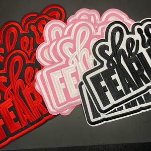 Pode incluir: Uma coleção de emblemas bordados com o texto "she is FEARLESS" numa fonte estilizada. Os emblemas estão disponíveis em três variações de cor distintas: vermelho com bordas pretas, rosa claro com bordas brancas e preto com bordas brancas.
