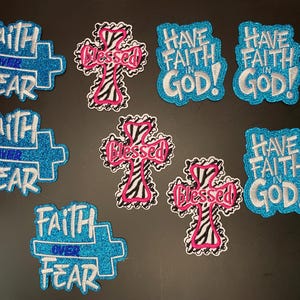 May include: A collection of embroidered patches featuring three blue glitter cross-shaped patches with the text Faith Over Fear, three cross-shaped patches with a black and white zebra print and pink Blessed text, and three blue glitter patches with Have Faith In God.