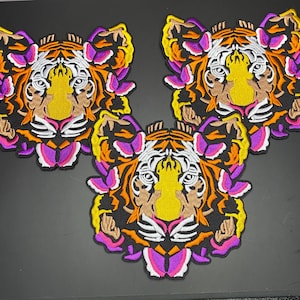 May include: Three embroidered tiger head patches featuring a vibrant color palette of orange, yellow, white, black, and magenta. Each patch displays a stylized tiger face with intricate thread work, detailed facial features, and bold outlines.