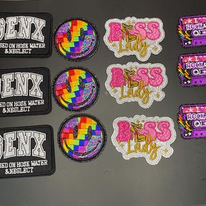 May include: A collection of embroidered patches including three rectangular black patches with silver text reading GENX RAISED ON HOSE WATER & NEGLECT, three circular rainbow checkered smiley faces, three pink and gold BOSS LADY patches, and three purple cassette tape patches with I LOVE ROCK & ROLL text.