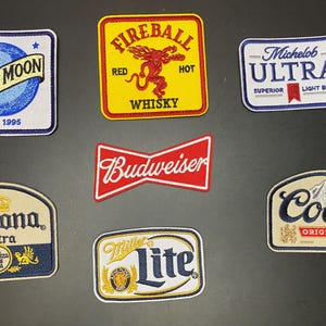 May include: A collection of seven embroidered patches featuring various beverage brand logos including Blue Moon, Fireball Whisky, Michelob Ultra, Budweiser, Corona Extra, Miller Lite, and Coors, each with distinct colors, shapes, and brand text.