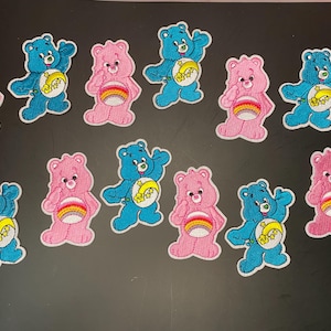 May include: A collection of twelve embroidered patches featuring cartoon bears in blue and pink. The blue bears display a yellow crescent moon and stars on their bellies, while the pink bears feature a colorful rainbow design on their bellies.