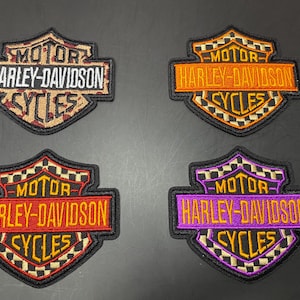 May include: Four shield-shaped embroidered patches featuring the Harley-Davidson Motor Cycles logo. One patch has a tan leopard print background, while the other three feature checkered patterns in orange, red, and purple color schemes respectively.