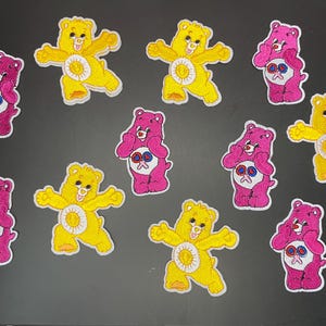 May include: A collection of ten embroidered patches featuring cartoon bears. Five patches are yellow bears with a sun emblem on their bellies, and five are pink bears with a heart-shaped emblem on their bellies. Each patch has a white border.