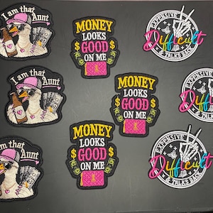 May include: A collection of nine embroidered patches featuring three distinct designs: a goose wearing a pink hat and sunglasses with the text I am that Aunt, a pink purse with the text Money Looks Good On Me, and a skeleton hand with the text Expensive & Difficult.
