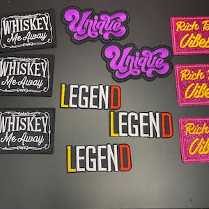 Peut inclure: Une collection de patchs brodés comprenant trois patchs rectangulaires noirs avec le texte blanc Whiskey Me Away, deux patchs en écriture cursive violette indiquant Unique, trois patchs rectangulaires à paillettes roses avec du texte jaune Rich Bitch Vibes et trois patchs avec le mot LEGEND.