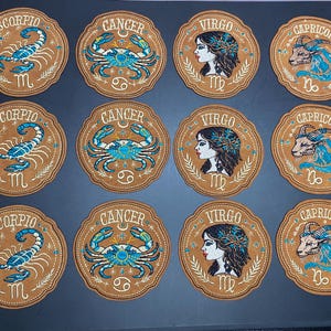 May include: A collection of twelve circular embroidered patches featuring four zodiac signs: Scorpio, Cancer, Virgo, and Capricorn. Each patch has a tan background with blue and white thread detailing, displaying the sign name, symbol, and illustration.